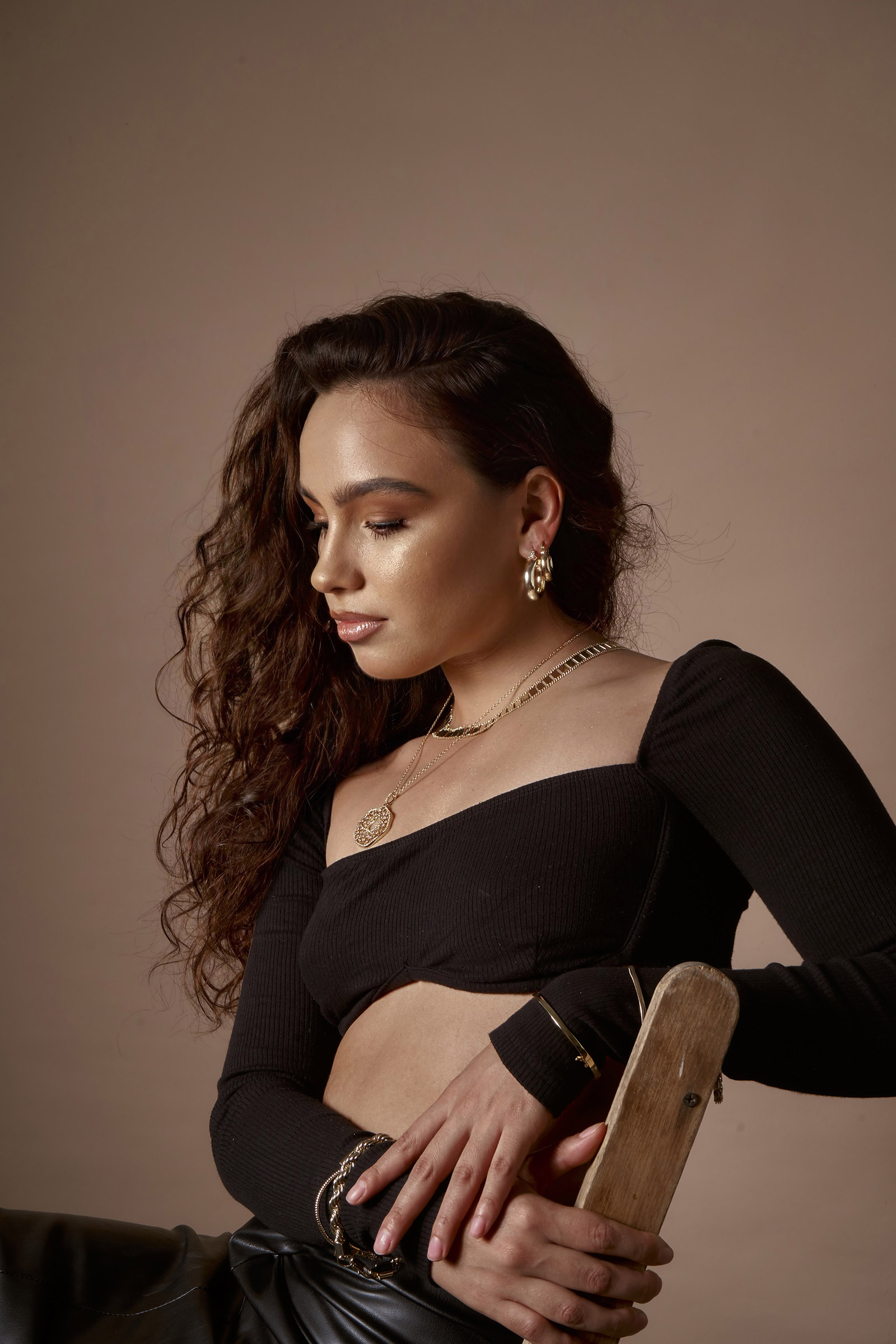 Abril Escalante a model with curly brown hair is captured in a side profile against a muted brown background. She wears a black off-shoulder top a gold chain necklace and matching gold earrings. Her fingers rest gently on a wooden object and she is adorned with several gold bracelets.